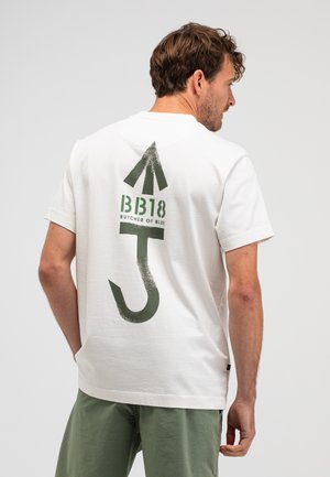 Man draagt een wit T-shirt met een groene grafische pijl, haakontwerp en de tekst "BB18 Butcher of Blue" op de achterkant, gecombineerd met groene korte broek.