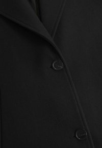 Manteau en laine noire avec un revers cranté, une rangée unique de boutons noirs, et une finition douce et texturée.
