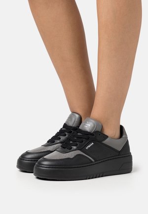 Sorte og grå lavtop-sneakers med sorte snørebånd, båret på bare fødder, med teksten "CPHGN" på siden og en tyk sål.