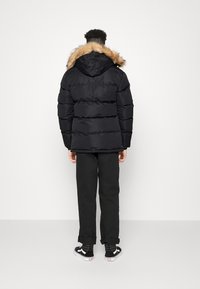 Mann trägt eine schwarze gepolsterte Parka mit einem mit Fell besetzten Kapuzen, schwarze Hosen und schwarze Sneakers in hoher Form, steht vor einer weißen Wand.