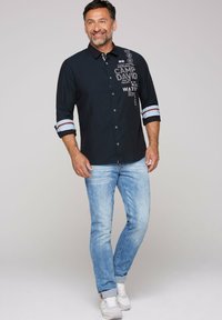 Camisa de botones navy con texto blanco y diseños gráficos, mangas enrolladas con acentos a rayas, combinada con jeans de color azul claro y zapatillas blancas.