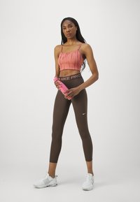 Rozā ribota topa ar plānām siksnām, kombinēts ar brūniem augsto vidukli legingiem ar melnu Nike Pro jostu. Modelis tur rozā ūdens pudeli.