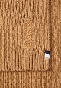 Stickad brun halsduk med ribbad textur. Har "BOSS"-logotyp i matchande brodyr och en randig etikett i svart, vitt och orange.