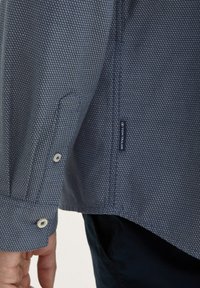 Camisa azul con un estampado, de tejido texturizado, que presenta un puño abotonado y una etiqueta cosida, exhibida en un brazo.