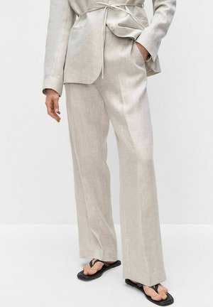 Persoon draagt een beige linnen kostuum met rechte broekspijpen en zwarte teenslippers, hand in zak, staand tegen een witte achtergrond.