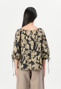 Blouse noire avec motif floral beige, design décolleté, manches bouffantes avec liens aux poignets, tissu fluide et coupe décontractée.