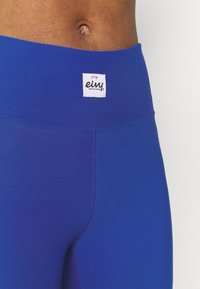 Modré legíny s vysokým pasem vyrobené z elastického materiálu. Mají plochý pas s bílou značkou a hladkou, rovnoměrnou texturu.