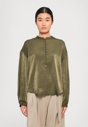 Femeie îmbrăcată cu o bluză de satin verde măslin, cu nasturi acoperiți pe față, asortată cu pantaloni largi bej cu talie înaltă.