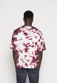 Camiseta de manga corta, teñida en anudado, de color blanca con patrones burdeos. Corte holgado, cuello redondo, material de algodón. Presenta diseños abstractos variados.