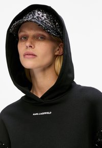 Young woman with freckles wearing black hoodie and sequined black cap, facing forward with neutral expression on white background.