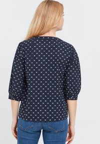 Blusa blu navy con motivi floreali bianchi, maniche a tre quarti e dettagli con bottoni ai polsini. Tessuto liscio con righe verticali.