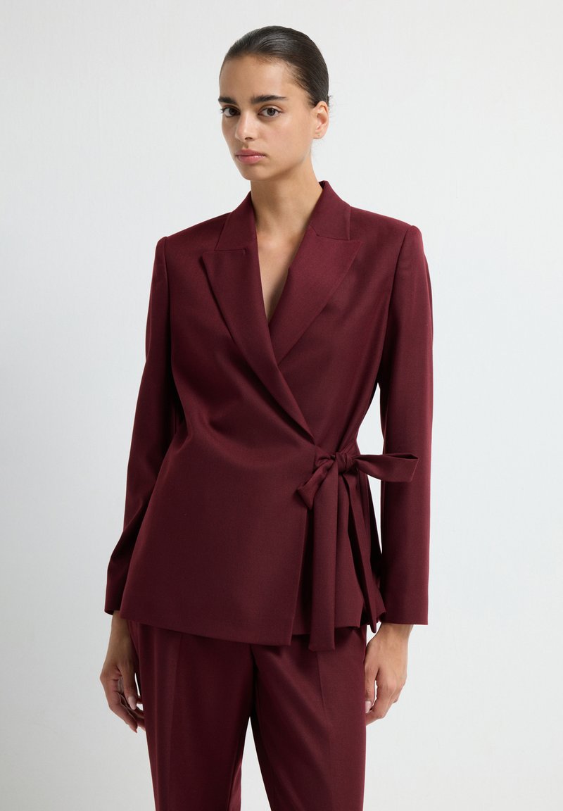 Blazer avvolgente bordeaux con maniche lunghe, dettaglio con cintura in vita, rever a incasso e tessuto liscio, abbinato a pantaloni coordinati.