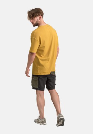 Korte mouwen, mosterdgele t-shirt met een ronde hals; gecombineerd met zwarte shorts met groene cargozakken en grijze sportschoenen.