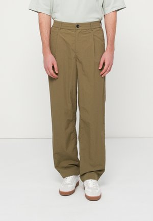 Άτομο που φορά χαλαρά ελαιδί παντελόνια και λευκά αθλητικά παπούτσια, στέκεται μπροστά σε απλό λευκό φόντο.