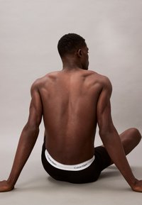 Schwarze Calvin Klein Unterwäsche mit einem weißen Logo-Bund. Das Modell sitzt und zeigt einen definierten Rücken sowie eine glatte Hauttextur vor einem neutralen Hintergrund.