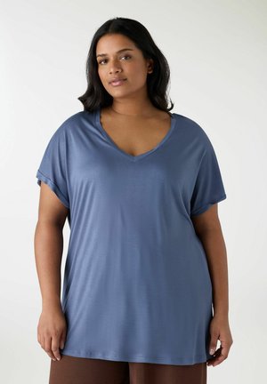 Donna con lunghi capelli scuri che indossa una maglia blu a maniche corte con scollo a V e pantaloni marroni, in piedi su uno sfondo bianco.