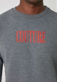 Sudadera gris con texto bordado en rojo que dice "COUTURE". Cuello redondo y textura suave, adecuada para el uso casual.
