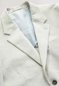 Veste de costume beige clair avec col à revers cranté, tissu légèrement texturé, boutonnière sur le revers gauche et bouton foncé près du bord inférieur.