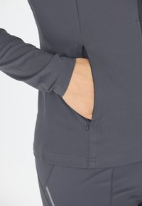 Grå træningsbluse med lange ærmer og en lynlåslomme. Stoffet har en glat tekstur, og den har diskrete stikninger.