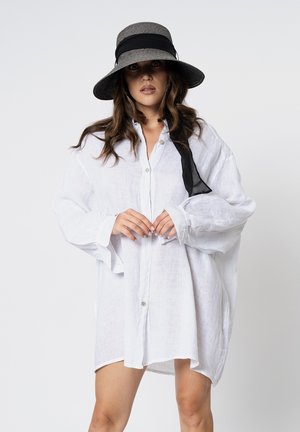 Donna che indossa una camicia bianca oversize con bottoni e un cappello a tesa larga bianco e nero, in piedi contro uno sfondo chiaro e semplice.