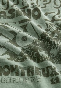 Grüner Stoff mit großen, fett gedruckten Texten in Dunkelbraun sowie verschiedenen Formen und Designs, die ein texturiertes Erscheinungsbild erzeugen.