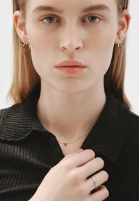 Collarín negro acanalado, collar de oro con una pequeña joya roja, anillo delicado en la mano, múltiples pendientes en las orejas, textura de piel suave.
