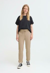 Zwart shirt met korte mouwen gecombineerd met beige broeken met een koord in de taille, gedragen met zwart-witte sneakers. Effen achtergrond.