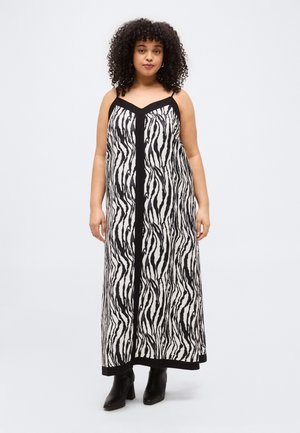 Kvinde med krøllet hår iført en sort og hvid zebra-stribet ærmeløs maxikjole og sorte ankelstøvler, stående mod en ensfarvet baggrund.