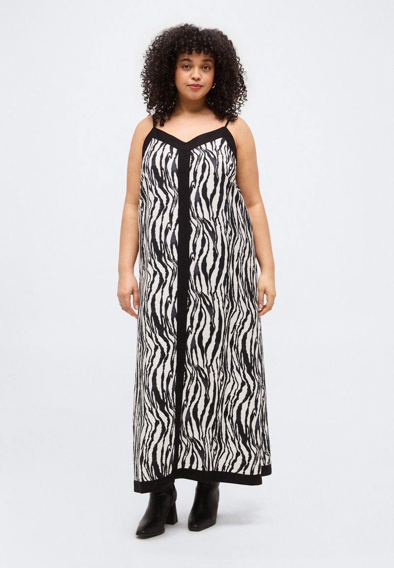 Γυναίκα με σγουρά μαλλιά, που φοράει μακρύ ασύμμετρο φόρεμα χωρίς μανίκια σε μαύρο και λευκό μοτίβο ζέβρας και μαύρες μποτάκι μέχρι τον αστράγαλο, στέκεται μπροστά σε ένα απλό φόντο.