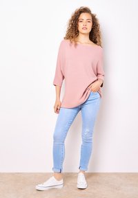 Hellrosa übergroßer Pullover mit weitem Ausschnitt, dreiviertellangen Ärmeln, kombiniert mit hellblauen Skinny-Jeans und weißen Sneakers.