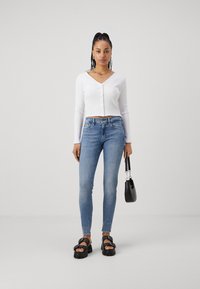 Camisola branca com nervuras, de mangas compridas e decote em V, combinada com jeans skinny azuis claros. Os acessórios incluem uma pequena bolsa preta e sandálias pretas chunky.
