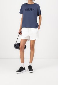 Marineblauwe katoenen T-shirt met "GANT" logo, gecombineerd met witte shorts, zwarte schoenen en een kleine zwarte tas. Eenvoudige, relaxte pasvorm.