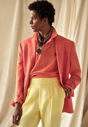 Homme portant un blazer corail, un polo corail, un pantalon jaune, un foulard à motif autour du cou, des lunettes de soleil et une montre, regardant sur le côté sur un fond beige.