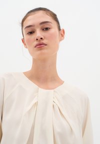 Blusa color crema con scollo tondo, caratterizzata da un dettaglio arricciato sul davanti e un tessuto morbido e leggero. Design semplice ed elegante.