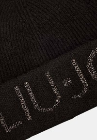 Beanie nera lavorata a maglia realizzata in morbido materiale, con una texture a coste e logo metallico argento dettagliato sul risvolto.