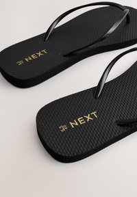 Tongs noires texturées avec fines brides et marquage doré "NEXT" avec détails de taille sur la semelle, présentées sur un fond clair.