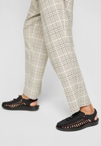 Zwarte gevlochten sandalen met een open achterkant, met een gestructureerde bovenkant en een gevoerde zool, gecombineerd met beige geruite broek.