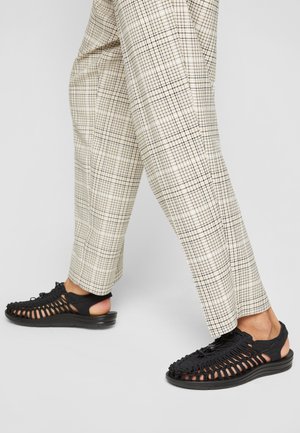 Zwarte open teen sandalen met een gevlochten bovenkant, rubberen zool en verstelbare veters. Gecombineerd met beige geruite broek voor contrast.