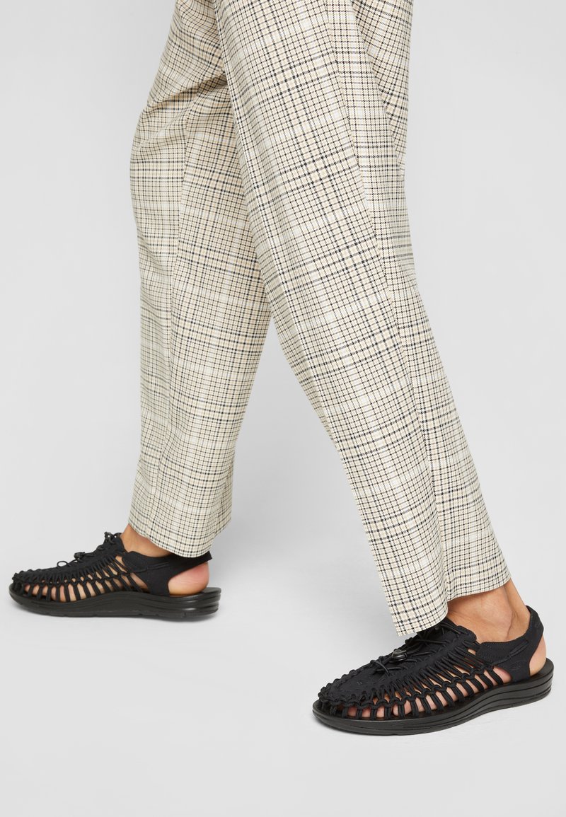 Zwarte gevlochten sandalen met een open achterkant, met een gestructureerde bovenkant en een gevoerde zool, gecombineerd met beige geruite broek.