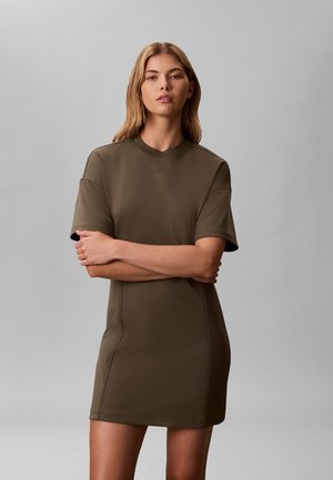 Joven mujer con cabello rubio largo, vestida con un vestido verde oliva de manga corta, de pie con los brazos cruzados frente a un fondo gris liso.