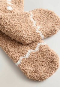 Hellbraune, flauschige Socken mit einer weichen Textur, die ein weißes Zickzackmuster aufweisen. Das Design beinhaltet abgerundete Kanten und ein dickes, gemütliches Material.