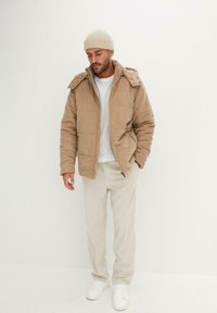 Giubbotto imbottito in corduroy beige con cappuccio, dotato di chiusura con zip e bottoni a pressione, indossato sopra una t-shirt bianca e pantaloni color crema, con sneaker bianche.