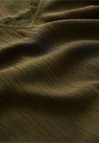 Olivgrön texturerad tyg med subtila åsar och en vågig yta, med delikat spetsdetaljering längs kanterna.