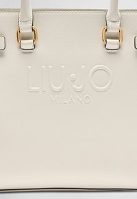 Balta tekstūruota pirkinių rankinė su įspaustu "LIU•JO MILANO" logotipu ir auksinės spalvos detalėmis. Turi tvirtas, reguliuojamas rankenas.