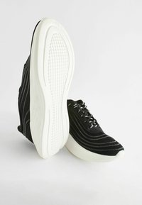 Black athletic shoes featuring a knit upper with wavy white stripes and white rubber soles. Laces are black with silver accents.