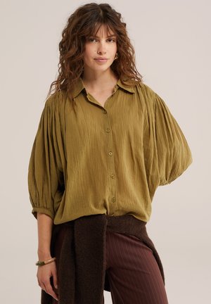 Femme aux cheveux bouclés portant un chemisier texturé vert olive à boutons avec des manches bouffantes et un pantalon marron foncé avec un pull noué à la taille.