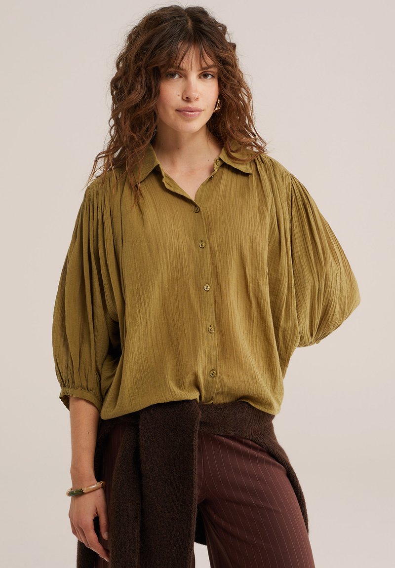Femme aux cheveux bouclés portant un chemisier texturé vert olive à boutons avec des manches bouffantes et un pantalon marron foncé avec un pull noué à la taille.