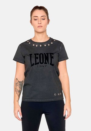 Frau trägt ein schwarzes "LEONE 1947" T-Shirt mit Metallösen am Kragen und Saum, steht vor einem einfarbigen, hellen Hintergrund.