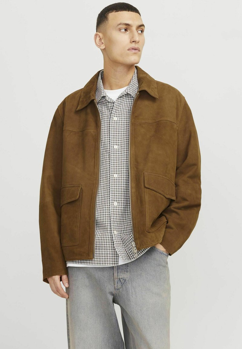 Brown suede jacket with a collared neckline, two front pockets, and a relaxed fit, layered over a checkered shirt. Gray jeans complete the look.
