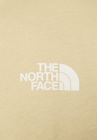 Béžová bavlněná tkanina s bílým logem "THE NORTH FACE" s obloukovým designem umístěným v dolním rohu. Texturovaný povrch.